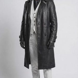 Pode incluir: Um sobretudo longo de couro preto usado sobre um colete branco, calças brancas, camisa preta e gravata preta. O traje inclui luvas de couro preto e sapatos de couro preto. A pessoa tem cabelo claro e usa óculos de sol escuros.