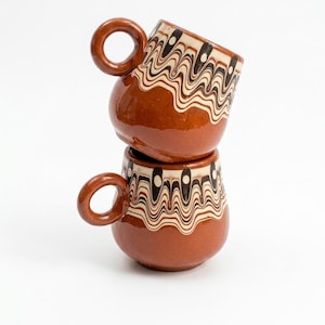 Puede incluir: Dos tazas de cerámica marrón apiladas. Cada taza tiene un cuerpo redondeado, un asa circular y una banda decorativa de líneas onduladas negras y blancas cerca del borde. Las tazas están sobre un fondo blanco liso.