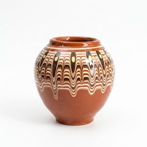Puede incluir: Un jarrón redondo de terracota con una base lisa de color marrón sólido y una sección superior decorativa adornada con un patrón ondulado y jaspeado en tonos marrón oscuro, crema y blanco, acentuado por pequeños puntos ovalados amarillos.