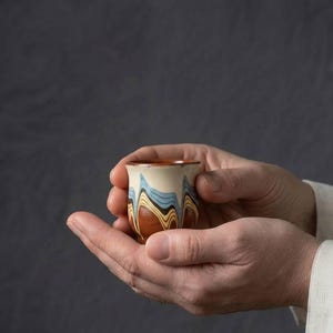 Puede incluir: Una pequeña taza de cerámica con un patrón jaspeado de bandas onduladas en crema, azul claro, marrón oscuro y amarillo. El recipiente tiene un borde acampanado y un cuerpo liso y redondeado, mostrando una técnica de arcilla estratificada.