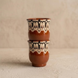 Puede incluir: Dos tazas de cerámica apiladas con un acabado liso y brillante. Las secciones inferiores son de un color marrón terracota sólido, mientras que los bordes superiores están decorados con un patrón ondulado y jaspeado en crema, marrón oscuro y negro.