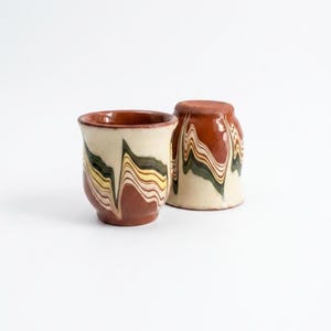 Puede incluir: Dos tazas de cerámica con base y borde marrón, y cuerpo color crema. Las tazas presentan un patrón ondulado en tonos marrón, amarillo y verde. Una taza está en posición vertical y la otra al revés.