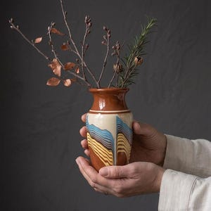 Puede incluir: Un pequeño jarrón de cerámica con borde acampanado y acabado liso y brillante. El cuerpo presenta un patrón jaspeado de líneas onduladas horizontales en tonos azul, amarillo y marrón. Varias ramas desnudas con pequeños brotes están colocadas dentro.