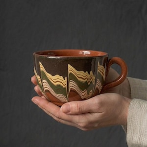 Puede incluir: Una taza de cerámica con un patrón jaspeado de remolinos en marrón oscuro, verde oliva y crema. El exterior tiene un acabado brillante, mientras que el interior y el asa curva revelan una base de arcilla de color terracota sin esmaltar.