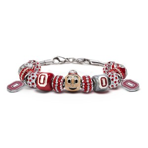 May include: A silver charm bracelet with red and white charms, including a charm with the Ohio State Buckeyes mascot, Brutus Buckeye. The bracelet also features charms with the letter "O" and red and white crystal beads.