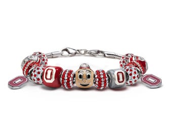 May include: A silver charm bracelet with red and white charms, including a charm with the Ohio State Buckeyes mascot, Brutus Buckeye. The bracelet also features charms with the letter "O" and red and white crystal beads.
