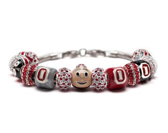 May include: A silver charm bracelet featuring various beads in red, black, and gray. Some beads are encrusted with red rhinestones, while others display the letter "O" or a cartoon character face. The bracelet is on a white background.