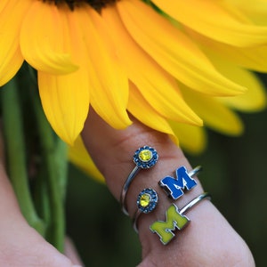 May include: Three silver rings with blue and yellow enamel. The rings feature a small yellow gem, the letter "M" and the letter "M" with a blue gem.