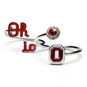May include: Set of three silver rings with red and white enamel. The rings feature the letters "OH", the word "io", and the letter "O" in a white and red design. One ring has a red rhinestone surrounded by clear rhinestones.