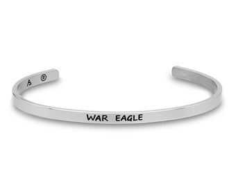 May include: A silver-colored cuff bracelet with the words "WAR EAGLE" engraved in black. The bracelet has a simple, open design and is likely made of stainless steel or a similar metal. The bracelet is a piece of jewelry.