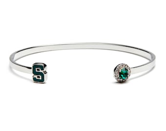 May include: A silver bangle bracelet featuring a green enamel letter 'S' and a green gemstone set in a halo of clear rhinestones.