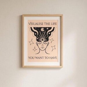 May include: A framed art print with a light wood frame. The artwork features a stylized illustration of a face with an eye in the center of the forehead, surrounded by stars and a crescent moon. The text reads "VISUALISE THE LIFE YOU WANT TO HAVE."