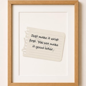 May include: A framed print featuring a torn piece of lined paper with the handwritten text: "Just make it exist first. You can make it good later." The frame is a light brown color, and the background is white.