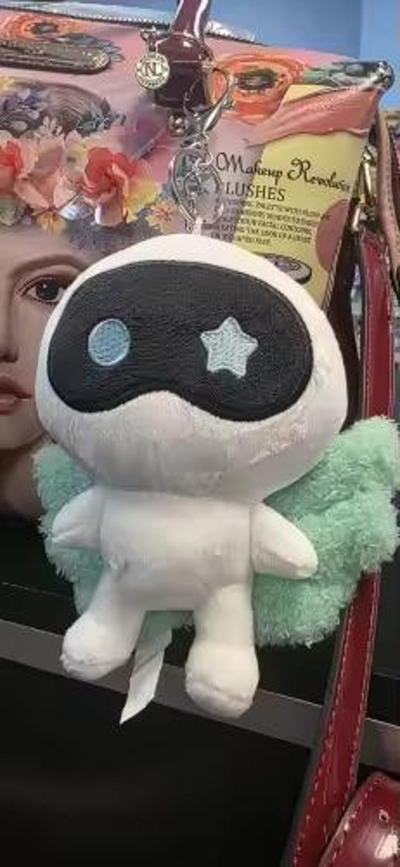May include: A white plush toy with a black mask, a blue circle, and a blue star. It has light green wings and a small white tag. The toy is attached to a silver keychain. The background includes a purse and a makeup product.