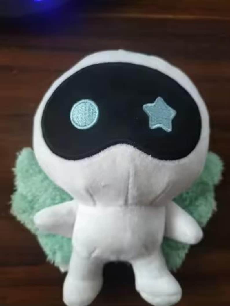 May include: A white plush toy with a black mask-like design and light blue embroidered details. The toy has a round embroidered circle and a star on the mask. It has small arms and green fluffy wings.