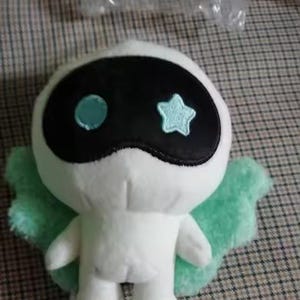 May include: A white plush toy with a black mask-like design on its face, featuring a blue circle and star. It has light green wings and is standing upright. The toy is on a checkered surface.