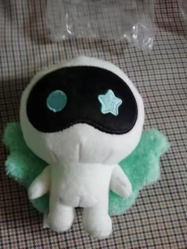 May include: A white plush toy with a black mask-like design on its face, featuring a blue circle and star. It has light green wings and is standing upright. The toy is on a checkered surface.