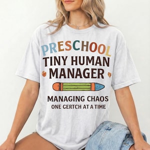 May include: A white short-sleeve t-shirt featuring the text "PRESCHOOL TINY HUMAN MANAGER" in multi-colored letters, a graphic of a green pencil, and the text "MANAGING CHAOS ONE CERTCH AT A TIME" in dark brown lettering on the front.