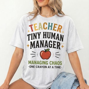 May include: A white short-sleeve t-shirt featuring the text TEACHER TINY HUMAN MANAGER in black and multicolored letters, a red apple graphic, and the text MANAGING CHAOS - ONE CRAYON AT A TIME in green and black, with small gold star accents.