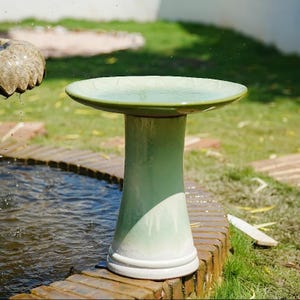 May include: A ceramic birdbath featuring a wide, shallow basin and a cylindrical pedestal base. The piece has a glossy glaze that transitions from a deep sage green at the top to a lighter, creamy white hue toward the bottom, with a smooth finish.