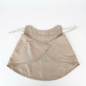 May include: A beige, wrap-style bib made of soft fabric. The bib has a rounded shape with a curved neckline and a drawstring closure. The fabric appears to be a natural fiber, possibly linen or cotton.