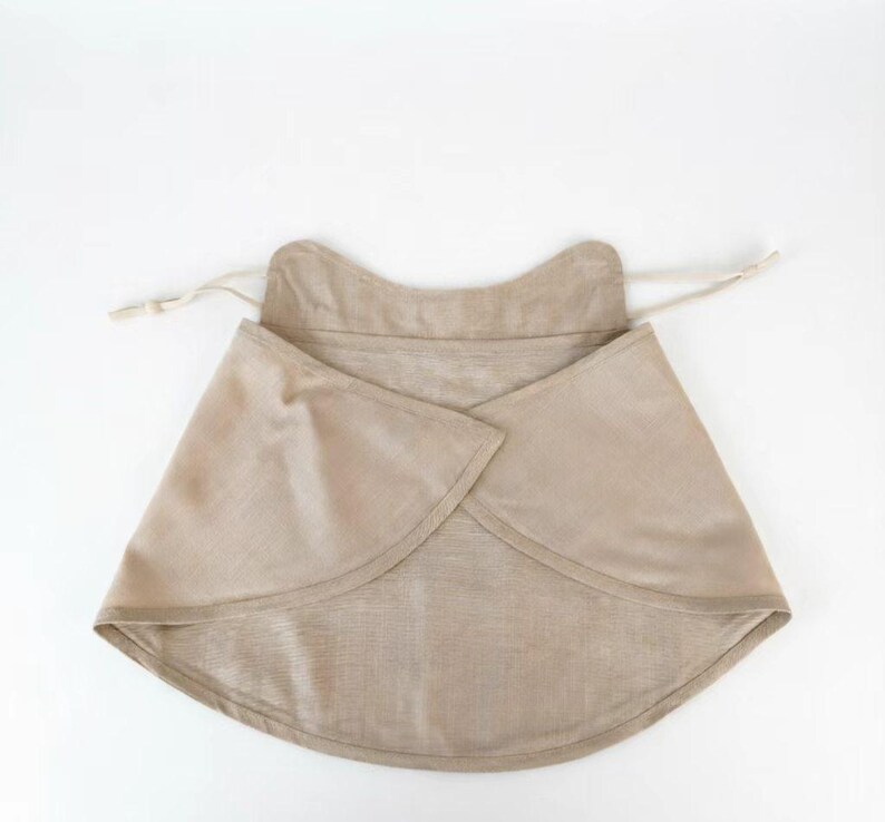 May include: A beige, wrap-style bib made of soft fabric. The bib has a rounded shape with a curved neckline and a drawstring closure. The fabric appears to be a natural fiber, possibly linen or cotton.