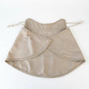 May include: A beige wrap-around garment with a curved hemline and adjustable ties. The fabric appears to be a lightweight linen or similar material. The garment is designed to be worn around the neck and shoulders.