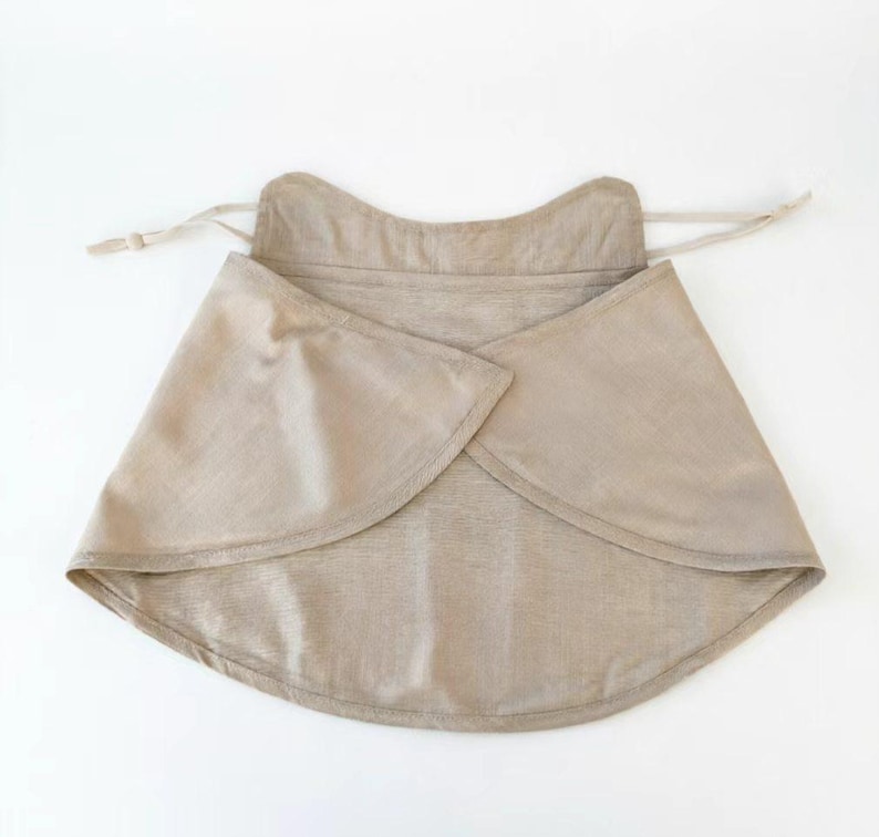 May include: A beige wrap-around garment with a curved hemline and adjustable ties. The fabric appears to be a lightweight linen or similar material. The garment is designed to be worn around the neck and shoulders.