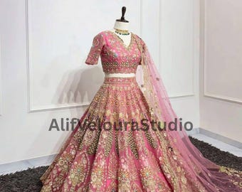 Peut inclure: Une lehenga rose brodée avec un haut assorti à manches courtes et un dupatta transparent. La lehenga présente des broderies complexes dorées et multicolores. Le texte "Alif Veloura Studio" est visible sur la jupe. La tenue est présentée sur un mannequin.