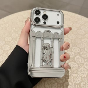 May include: A white smartphone case featuring a metallic silver finish. The design includes a raised relief of a cherub figure positioned between three vertical columns, with a metallic silver frame surrounding the camera lens cutout area.