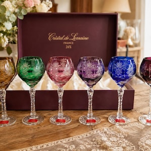 Peut inclure: Un ensemble de six verres à vin en cristal multicolore avec des gravures florales complexes et des pieds facettés, présentés dans une boîte violet foncé marquée Cristal de Lorraine France 24%. Chaque verre présente une teinte distincte : ambre, vert, rose, violet, bleu et rouge foncé.