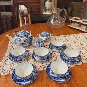 Peut inclure: Service à thé vintage avec un motif floral bleu et blanc. L'ensemble comprend une théière, un sucrier, une crémière et six tasses avec soucoupes. Le service est présenté sur une table en bois avec un napperon en dentelle.
