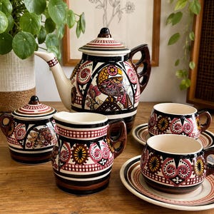 Peut inclure: Un service à thé en céramique comprenant une théière, un sucrier avec couvercle, un pot à lait et deux tasses avec soucoupes assorties. Chaque pièce est ornée de motifs géométriques et floraux complexes en noir, blanc, rouge et jaune.