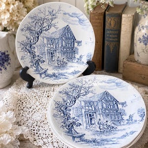 Peut inclure: Deux assiettes en céramique blanche avec un motif bleu. Les assiettes présentent une illustration détaillée d'un bâtiment avec des personnes et des arbres. Les assiettes sont présentées sur un napperon en dentelle blanche.