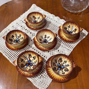 Peut inclure: Ensemble de six bols et soucoupes en céramique vintage. Chaque pièce présente un motif floral marron et jaune avec des touches de bleu et d'orange. Les bols ont de petites anses et sont posés sur une nappe en dentelle blanche.