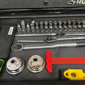 May include: A drawer filled with various tools, including sockets, ratchets, a torque wrench, oil filter wrenches, a tape measure, and a level. The tools are arranged neatly in a black drawer, with a green and black tape measure on the left and a yellow level on the right.