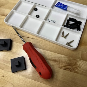 May include: A white divided tray filled with small hardware items, including screws, black plastic pieces, and glue. A red screwdriver and two black square objects are also present on a wooden surface.