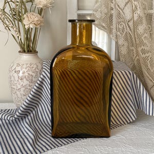 May include: A rectangular amber glass bottle with a dark rim. The bottle has a striped pattern and is displayed on a striped fabric. A white vase with floral designs and dried flowers are in the background.