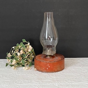 Puede incluir: Una lámpara de aceite vintage con una chimenea de vidrio transparente y una base de metal roja. La lámpara está sobre una superficie blanca.