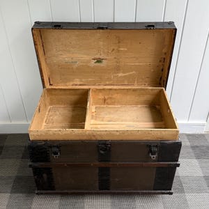 Puede incluir: Un ba&uacute;l de madera antiguo abierto, que revela un interior dividido. El exterior es marr&oacute;n oscuro con detalles y cierres met&aacute;licos. El ba&uacute;l est&aacute; sobre un suelo a cuadros. Dimensiones: aproximadamente 90 cm de largo.