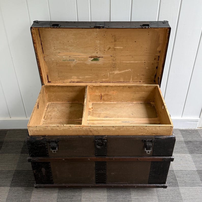 Puede incluir: Un ba&uacute;l de madera antiguo abierto, que revela un interior dividido. El exterior es marr&oacute;n oscuro con detalles y cierres met&aacute;licos. El ba&uacute;l est&aacute; sobre un suelo a cuadros. Dimensiones: aproximadamente 90 cm de largo.