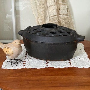 May include: A black cast iron oval pot with a matching lid featuring a lattice-style cutout pattern and side handles. Beside the pot sits a small, light-coloured ceramic bird figurine with textured wings and thin, dark metal wire legs.