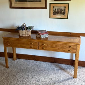 May include: A rectangular wooden console table with a light brown finish, featuring four drawers with small round brass knobs. A woven wicker basket filled with artificial flowers and a stack of three hardcover books sit on the surface.