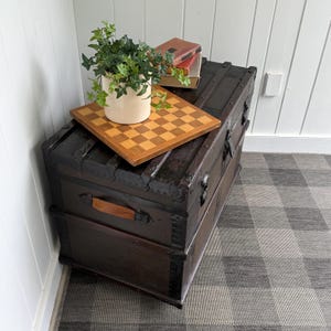 Puede incluir: Un ba&uacute;l antiguo marr&oacute;n oscuro con detalles met&aacute;licos y un asa de cuero. Un tablero de ajedrez de madera, una planta en maceta y dos libros descansan encima. El ba&uacute;l est&aacute; sobre una alfombra a cuadros gris y blanca, cerca de una pared blanca.