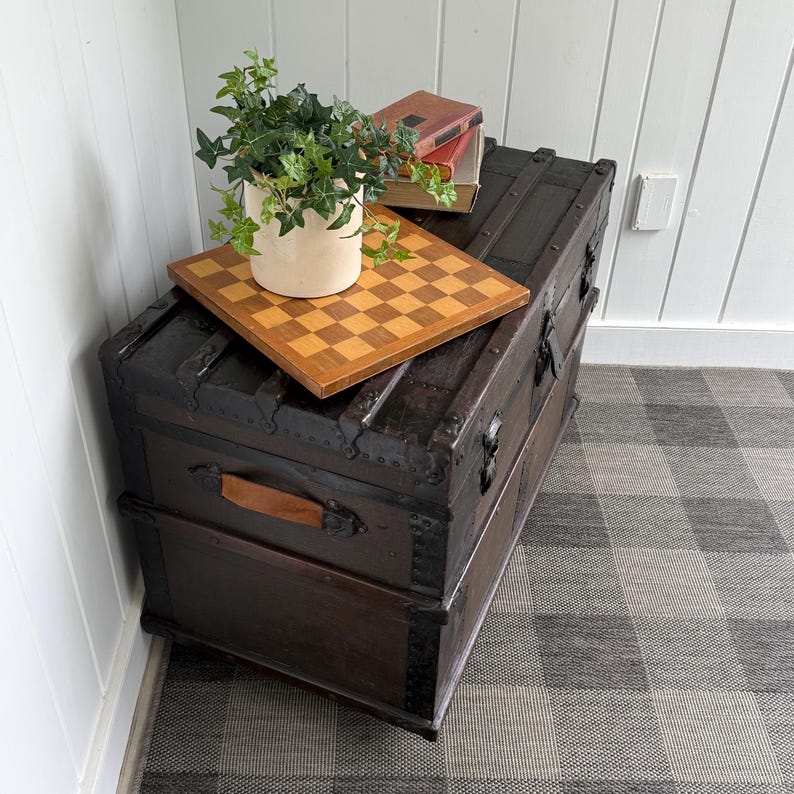 Puede incluir: Un ba&uacute;l antiguo marr&oacute;n oscuro con detalles met&aacute;licos y un asa de cuero. Un tablero de ajedrez de madera, una planta en maceta y dos libros descansan encima. El ba&uacute;l est&aacute; sobre una alfombra a cuadros gris y blanca, cerca de una pared blanca.