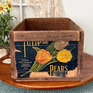 May include: Aged wooden fruit crate with a distressed label. The label displays an orange tulip and a yellow pear on a dark blue backdrop. Text on the label reads "TULIP BRAND PEARS UNITED STATES OF" and "Perham Fruit Company Yakima, Washington, U.S.A."