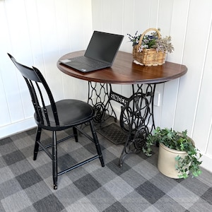 May include: A dark wood table with a black laptop, supported by a vintage Singer sewing machine base. A black chair sits to the left. A small basket of flowers and a potted plant are also present. The floor has a gray and white checkered pattern.