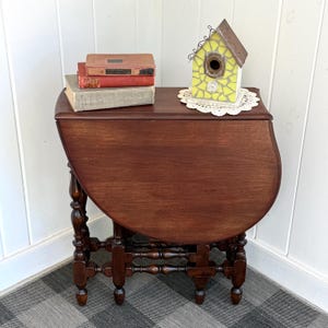 May include: A dark brown wooden drop-leaf table with turned legs, featuring a stack of three vintage books and a yellow mosaic birdhouse on a white lace doily placed on the tabletop surface. The table has a smooth, curved edge design.