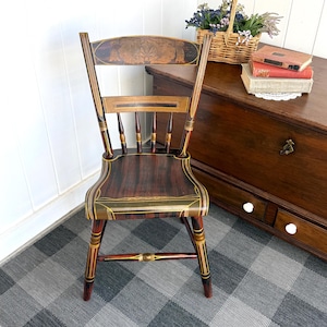 May include: An antique wooden chair with a dark brown seat and back, featuring decorative gold and red accents. The chair has a curved seat and back with a detailed design on the top. The legs are painted with red, gold, and black details.