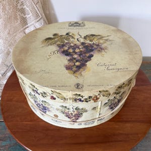 May include: Round decorative box with a vintage design. The lid features a cluster of purple grapes and the words "Cabernet Sauvignon." The box has a cream-colored background with a rope handle and grape illustrations around the sides.